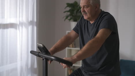 middle-aged-man-is-training-alone-at-home-using-modern-stationary-bike-for-workout-caring-about-health-keeping-fit-in-old-age